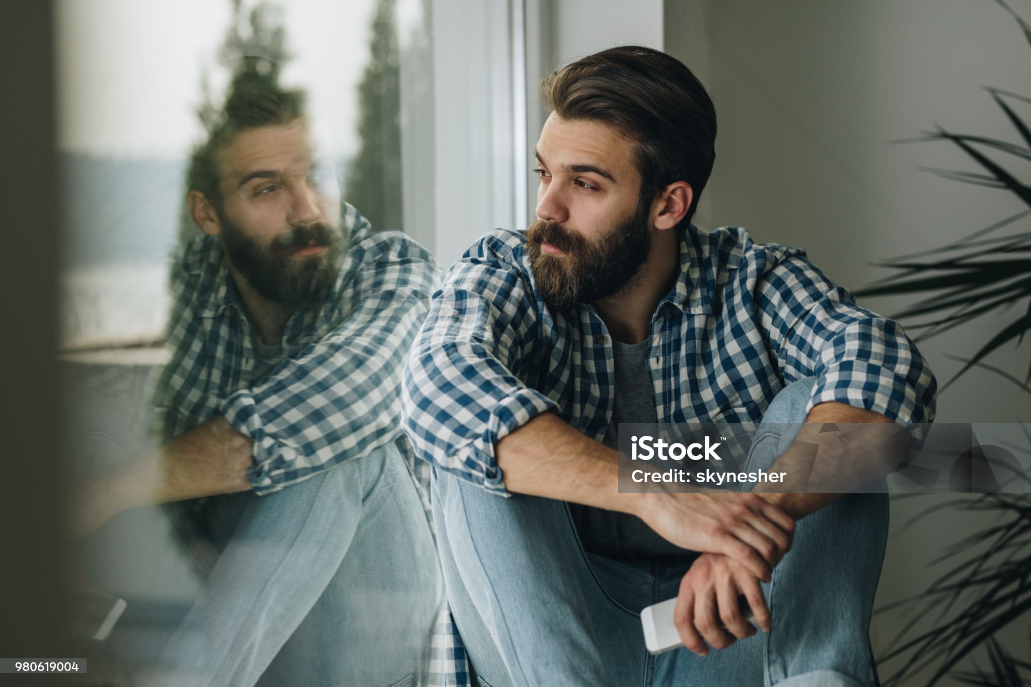 Young pensive man sitting by the window sill and looking through it. Young pensive man sitting by the window sill and looking through it.